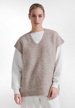 Kvinde med lyst hår iført en beige, blød, ærmeløs sweatervest over en hvid, langærmet top, stående mod en ensfarvet hvid baggrund.
