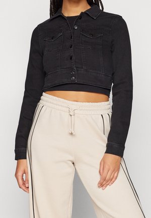 Donna che indossa una giacca corta di jeans nera sopra una maglia nera, abbinata a pantaloni della tuta beige a vita alta con strisce nere laterali e coulisse.
