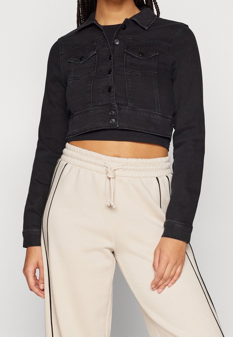 Femme portant une veste courte en denim noir sur un top noir, associée à un pantalon de jogging beige taille haute avec des bandes noires sur les côtés et un cordon de serrage.