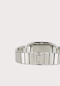 Reloj de acero inoxidable plateado con una cara rectangular y una pulsera segmentada. Presenta un cierre con un logo y un fondo texturizado.