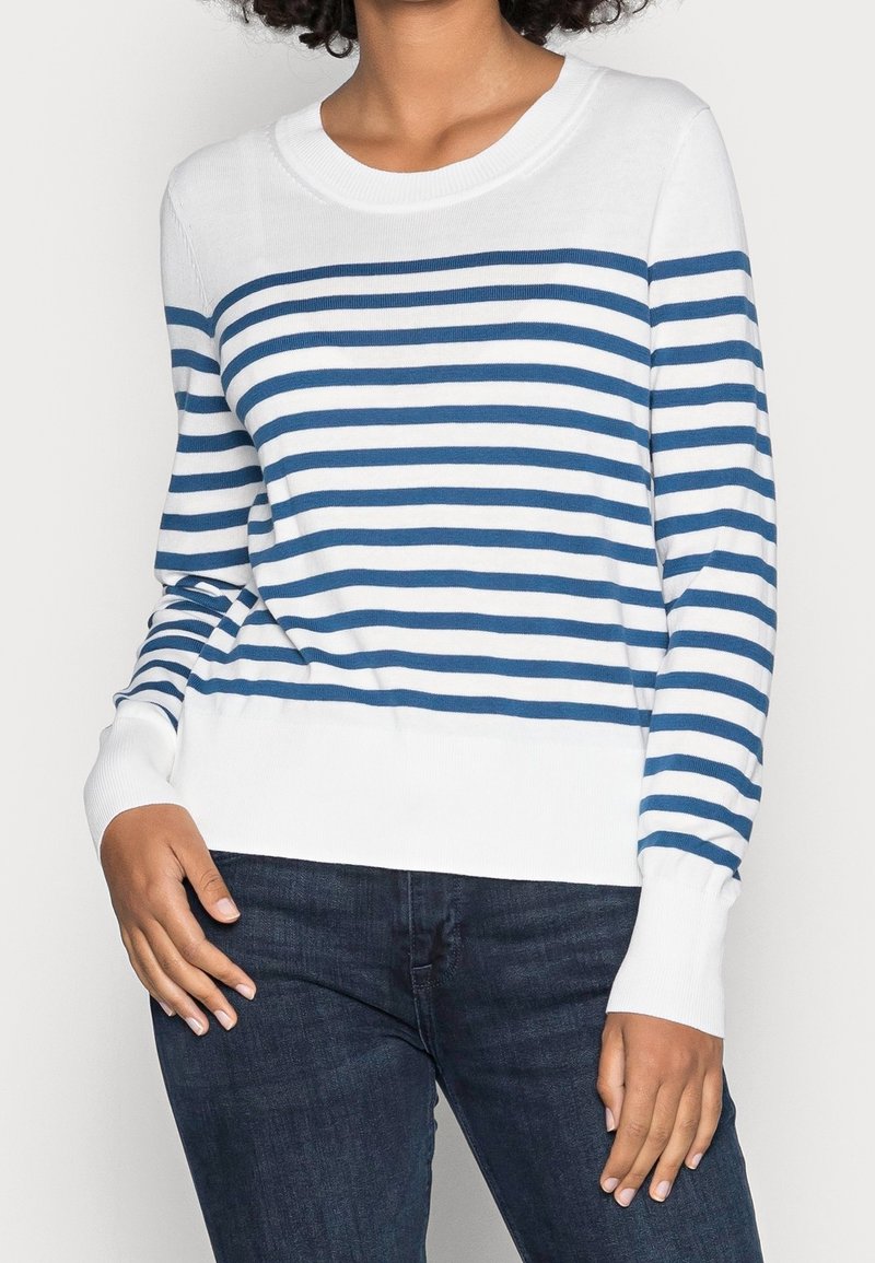 Femme portant un pull blanc à rayures bleues horizontales et un jean bleu foncé, debout avec une main sur la hanche.