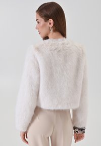 Weiße Fake-Fell-Cropped-Jacke mit weicher Textur, versehen mit langen Ärmeln und gemusterten Manschetten. Einfaches Design mit minimalistischem Finish.