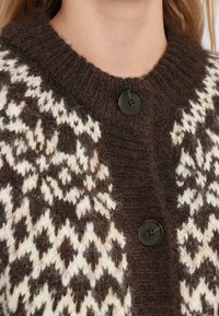 Brown and cream knitted cardigan with geometric patterns, featuring a high collar and a large dark button at the neck. Soft texture.