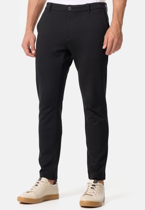 Schwarze, enganliegende Hose mit glatter Textur, ausgestattet mit einem Reißverschluss und einem Knopfverschluss. Kombiniert mit beigen Sneakern, die einen kontrastreichen Look zeigen.