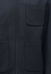 Σκούρο μπλε μπουφάν με λεία υφή, δύο μεγάλες τσέπες και κεντρικό φερμουάρ με ορατή λεπτομέρεια ραφής.