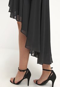 Jambes portant une robe noire asymétrique en dentelle et des sandales à talons hauts avec ankle-strap noires sur un fond blanc.
