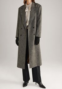 Cappotto grigio in tweed con davanti doppiopetto, rever a punte e tasche laterali. Abbinato a una blusa in satin, pantaloni neri e guanti.