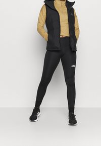 Černá péřová vesta přes béžové tričko s dlouhým rukávem, doplněná černými legínami a černými teniskami. Výrazné loga na vestě a legínách.