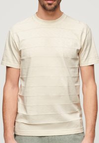 Camiseta de rayas beige con mangas cortas, fabricada en una tela suave. Presenta un escote redondo y sutiles líneas horizontales texturizadas.