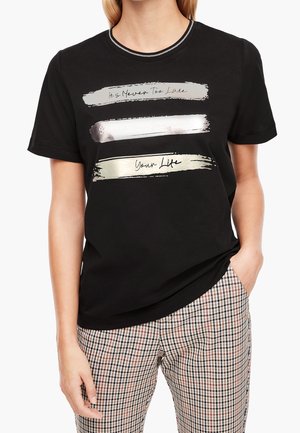 Kvinde iført en sort t-shirt med metallisk penselstrøgstekst "It's Never Too Late Your Life" og ternede bukser, hånd i lommen på hvid baggrund.