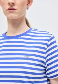 Γυναίκα που φοράει μπλε και λευκό T-shirt με οριζόντιες ρίγες και μικρό κεντημένο λογότυπο στο στήθος, μπροστά σε απλό ανοιχτό φόντο.