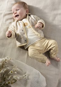 Beige baby-outfit met een shirt met bloemmotief en bijpassende broek, van zacht katoen met ronde randen. Liggend op een gestructureerde lichte achtergrond.