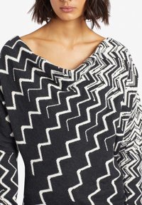 Kobieta ubrana w sweter w czarno-białym wzorze w zygzaki, z szerokim, luźno opadającym dekoltem i długimi rękawami.