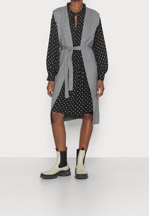 Femme portant une robe noire à pois, un gilet sans manches gris ceinturé, et des bottines crème et noires, debout sur un fond blanc.