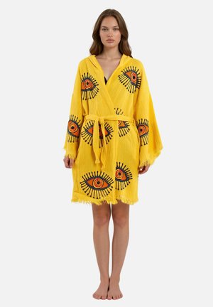 Donna che indossa un accappatoio giallo con cintura annodata, decorato con grandi disegni ricamati di occhi arancioni e neri, in piedi scalza.