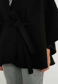 Capa de mezcla de lana negra con un acabado texturizado, que presenta un detalle de cintura atada y un dobladillo fluido. Acompañada de unos vaqueros grises para proporcionar contraste.