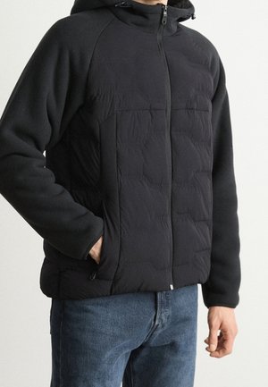 Mand iført sort hættetrøje med quiltet jakke med fleece-ærmer og blå jeans, en hånd i jakkelommen, står mod en ensfarvet baggrund.