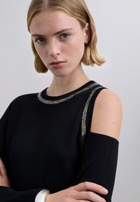 Suéter negro de punto con diseño de hombros descubiertos y ribete metálico plateado a lo largo del escote y las mangas. Textura suave, ajuste casual.