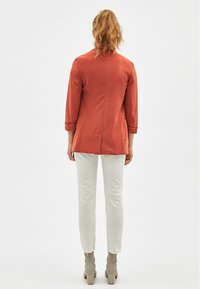 Bordový blazer se šitým střihem, dlouhými rukávy a jedním rozparkem vzadu, kombinovaný s bílými úzkými kalhotami a vzorovanými béžovými kotníkovými botami.