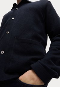 Dunkelblauer Cardigan aus einer weichen Wollmischung, mit Knopfverschluss, einer Tasche auf der linken Seite und gerippten Bündchen. Klassisches und minimales Design.
