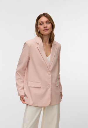 Femme portant un blazer oversize rose pâle et un pantalon blanc, debout devant un fond clair uni.
