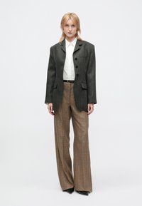 Vrouw met blond haar in een donkere streepjesblazer, witte shirt, bruine geruite wijde broek en zwarte puntige schoenen, staand op een witte achtergrond.