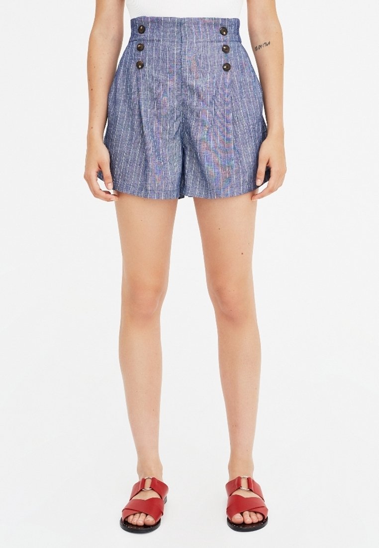 Shorts taille haute rayés bleu et blanc avec deux grands boutons à l'avant, des plis et une coupe ajustée. Associés à des sandales à glissière rouges.