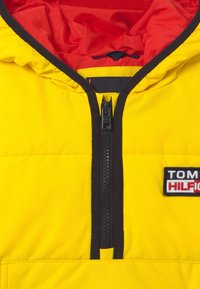 Żółta kurtka puchowa z czerwoną podszewką, czarnym zamkiem błyskawicznym i naszywką z logo. Posiada pikowaną teksturę oraz kaptur z elastycznym sznurkiem.