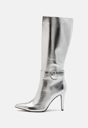 Bota plateada hasta la rodilla, de cuero suave y brillante, con punta estrecha y tacón delgado. Cuenta con una correa decorativa con hebilla alrededor del tobillo.