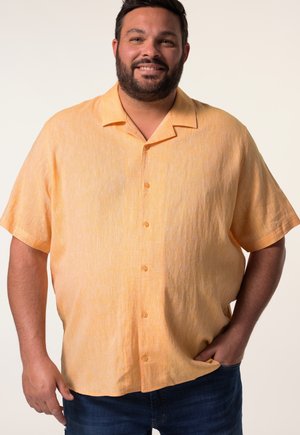 Mann mit Bart lächelnd, trägt ein kurzärmeliges orangefarbenes Hemd mit Knöpfen und dunkelblaue Jeans, steht mit einer Hand in der Tasche.