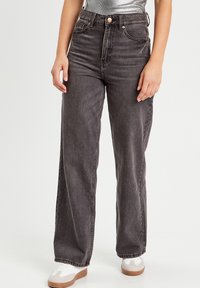 Jeans de tiro alto y pierna ancha en denim gris oscuro con un ligero desvanecimiento. Presentan un cierre de botón clásico y trabillas para cinturón. Combinados con zapatos blancos.