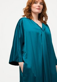 Tyrkysový saténový župan s V-výstřihem, krátkými křídly a uzlovým detailem na přední straně; má postranní kapsy a hladkou texturu.