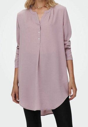 Vrouw draagt een losse, lange mouw, licht mauve tuniek met gedeeltelijke knopenlijst, gecombineerd met zwarte legging, staand tegen een witte achtergrond.
