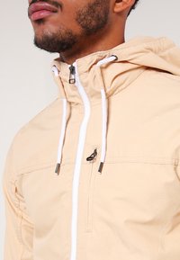 Peach-colored lightweight jacket with a zippered front, white drawstrings, and a hood. Features a chest pocket and smooth texture.