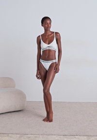 Witte beha- en bikinitopset met een gemerkte elastische band. Zacht, rekbaar materiaal met een eenvoudig ontwerp. Model poseert voor een neutrale achtergrond.