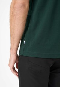 T-shirt di cotone verde scuro visibile di lato, con un'etichetta con accenti blu e bianchi all'orlo e pantaloni neri con tasca posterior.