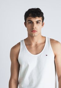 Weißes Baumwoll-Tanktop mit rundem Ausschnitt und schmalen Trägern, mit einem kleinen Logo auf der linken Brustseite. Sanfte Textur.