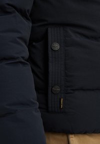 Nawigacyjna kurtka puchowa z teksturowanej tkaniny, wyraźnym stitchingiem i dwoma czarnymi guzikami z logo. Pomarańczowy detal metki na szwie.