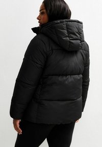Mujer con cabello largo y oscuro, vistiendo una chaqueta de plumas negra, vista desde atrás, de pie contra un fondo blanco liso.