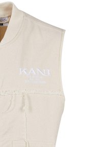 Beige ermeløs vest i bomull med ribbestrikket krage. Har frontal søm og brodert "KANI"-logo i hvit. Enkelt design.