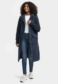 Marineblauwe lange jas met textuur gebreide mouwen, voorzakken, ritsluiting en verstelbare capuchon, gecombineerd met blauwe jeans en lichte sneakers.