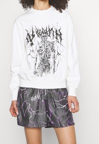 Vit sweatshirt med en grafisk skelettdesign, tillsammans med grå shorts med ett lila blixtmönster. Fleece-material, avslappnad passform.