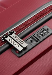 Maleta rígida roja en primer plano que presenta una superficie texturizada, cremalleras dobles con la marca "MODO" y un candado de combinación en el costado.