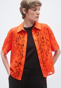 Fel oranje kanten T-shirt met korte mouwen en een ronde kraag, knopen aan de voorkant, gedragen over een zwart topje, met een losse pasvorm.