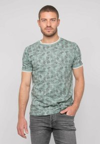 T-shirt vert clair à manches courtes avec un imprimé de feuilles noires, encolure et manches blanches contrastées, associé à un jean gris.