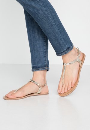 Füße in metallisch goldenen T-Riemchen-Sandalen mit flacher Sohle, kombiniert mit blauen Jeans, stehend auf einem hellen Boden.