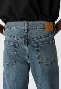 Person wearing blue denim jeans with back pockets and a brown leather patch labeled "WEEKDAY," paired with a black top.