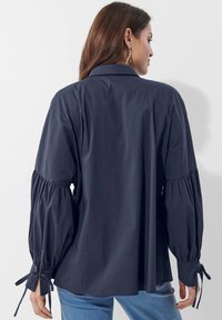 Marineblaue Bluse mit langen, gepufften Ärmeln, die mit gerafften Details und Bindebändern an den Handgelenken versehen ist; aus einem glatten Stoff gefertigt, lockere Passform.