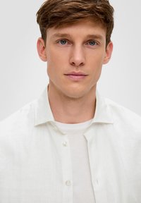 Witte linnen overhemd met knopen, gedragen over een wit T-shirt. Het overhemd heeft een relaxte kraag en een subtiele textuur. Glad huid en licht haar.