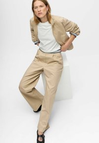Beige katoenen jas met elastische manchetten, wit t-shirt met zwarte rand, en beige broek met wijde pijpen. Zwarte schoenen met een strapdetail.